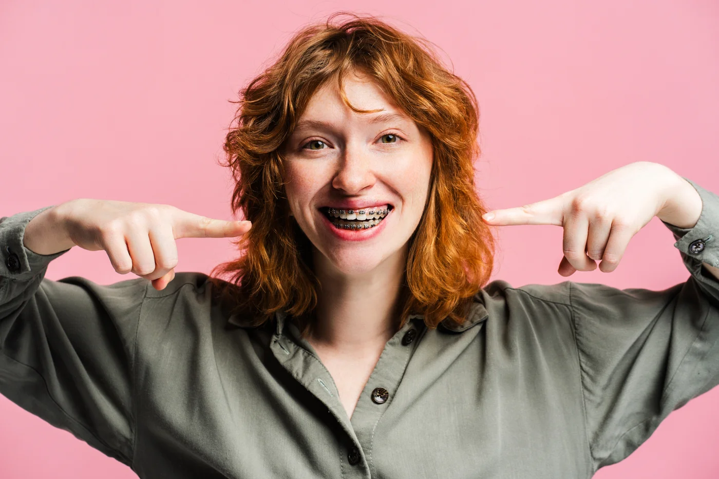Smiling person pointing to their braces against a pink background
