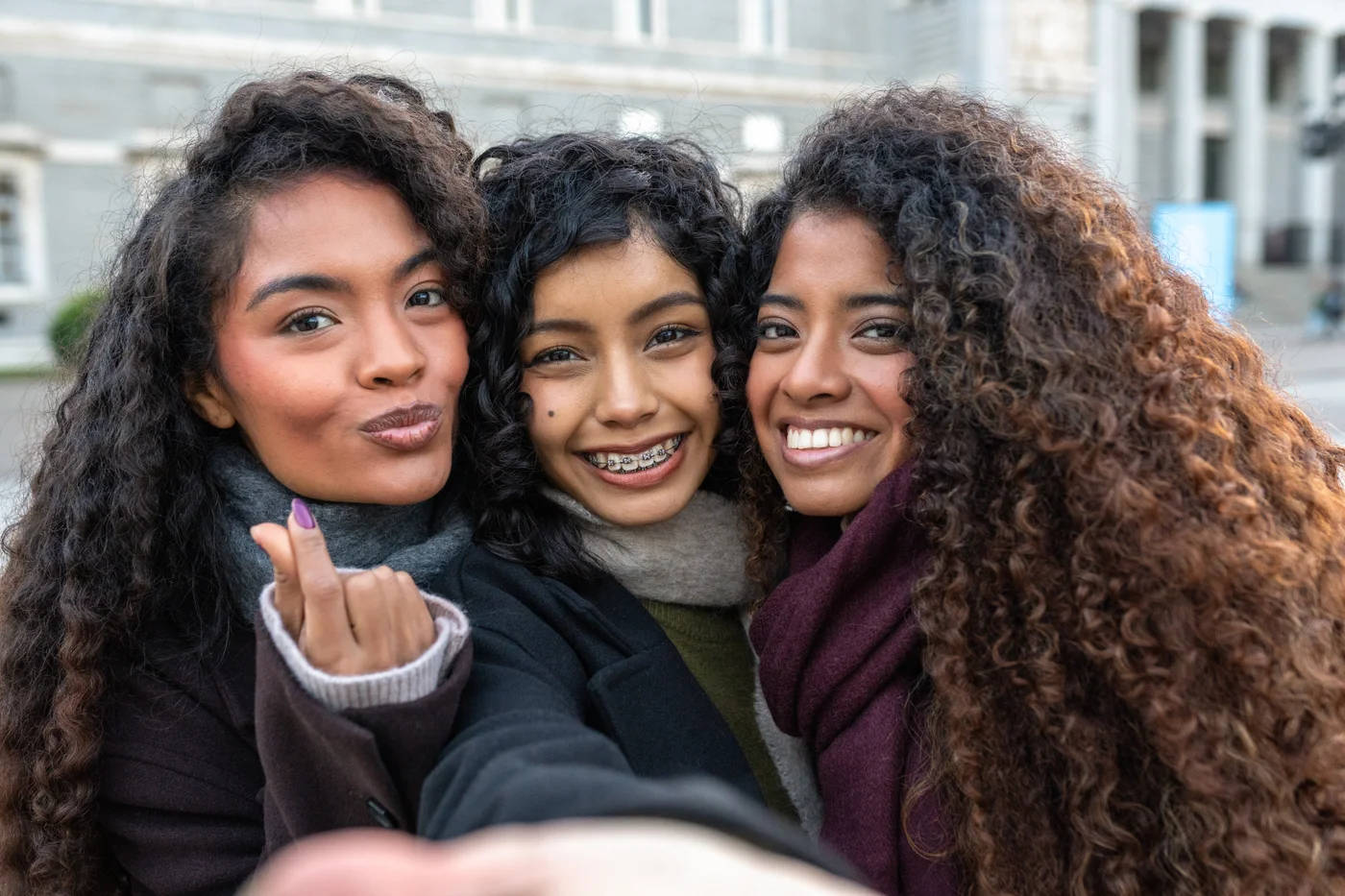 Three friends smiling together outdoors, with braces visible on the person in the center